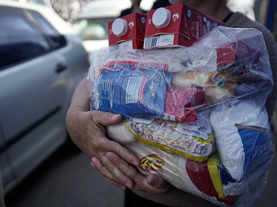 Mãos segurando uma sacola com alimentos de uma cesta básica