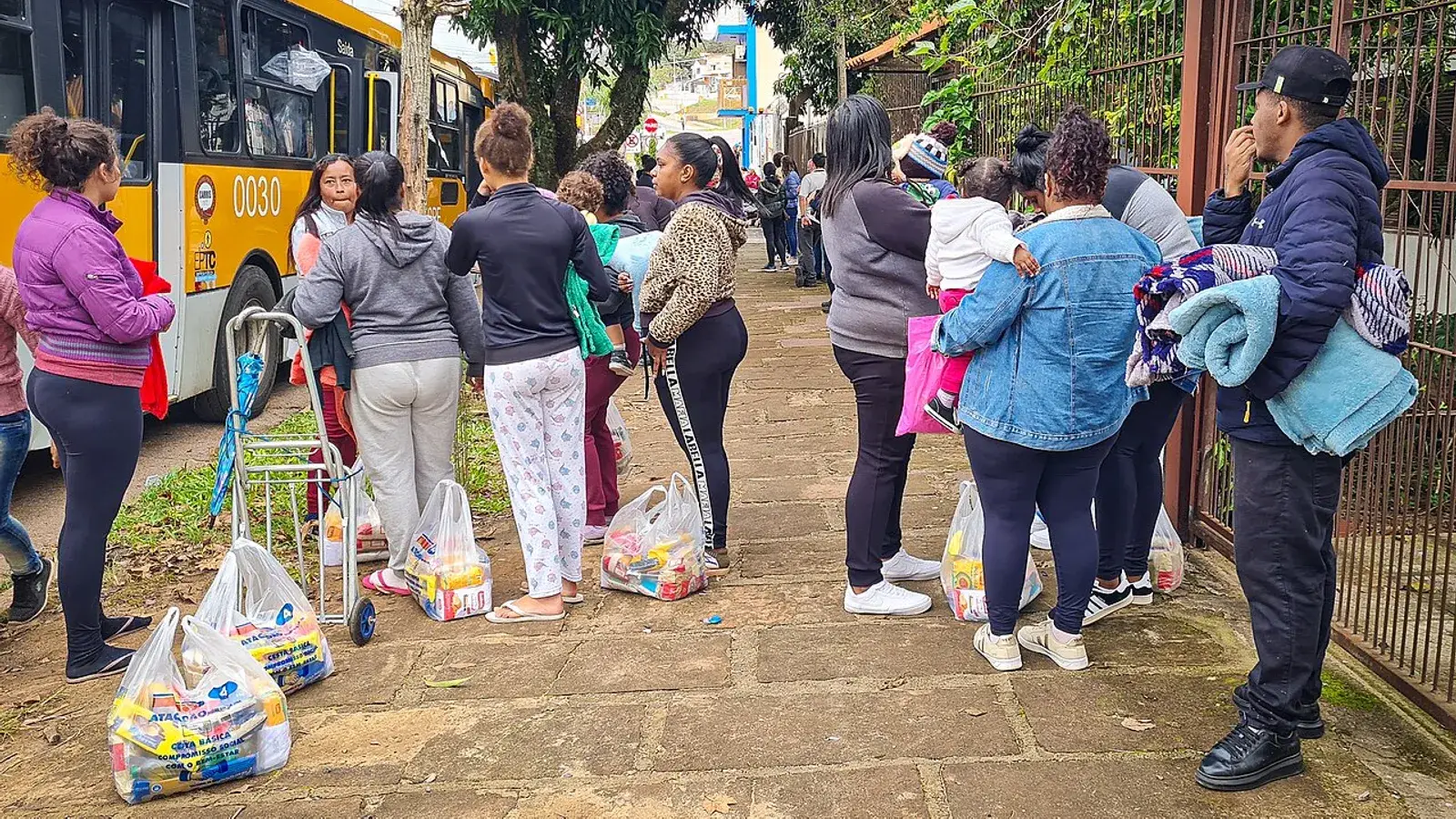 Famílias em fila com sacolas de alimentos ao lado de um ônibus