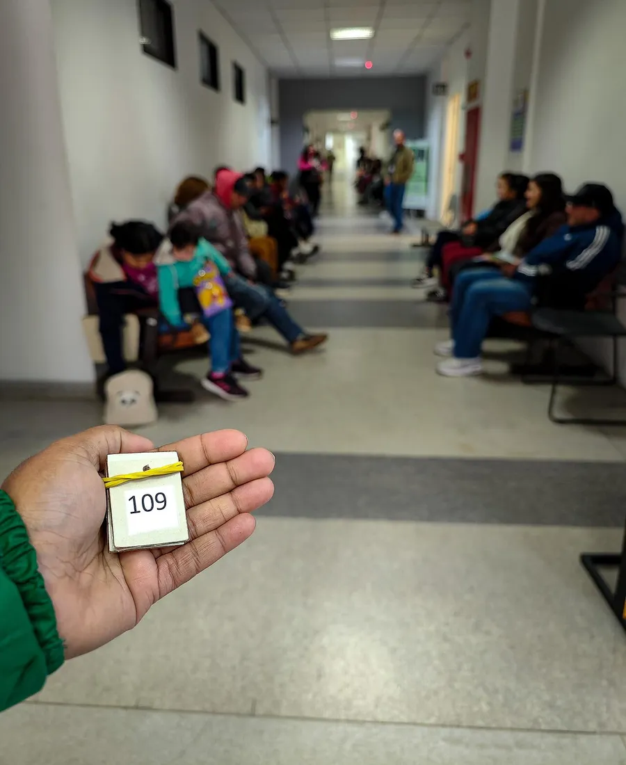 Mão segurando a senha de número 109 num corredor de espera com pessoas sentadas